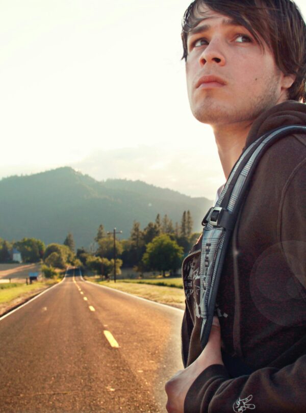 A young man with a backpack looks thoughtful on a scenic road with mountains at sunset.